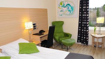 Een hotelkamer met een bed, groene kussens, een bureau met telefoon, een groene stoel, een klein tafeltje met lamp en een schilderij aan de muur.