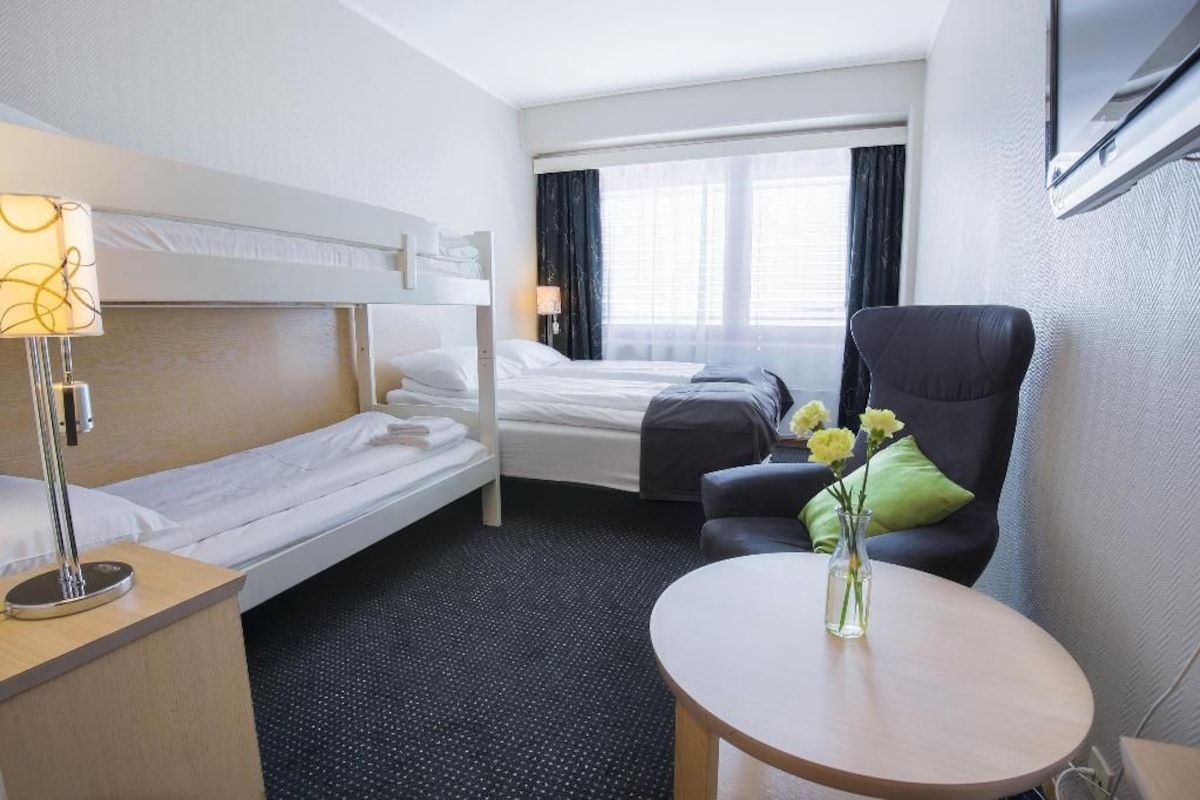 Habitación de hotel con literas, una cama individual, un escritorio con lámpara, una silla, una mesa redonda y un jarrón con flores.
