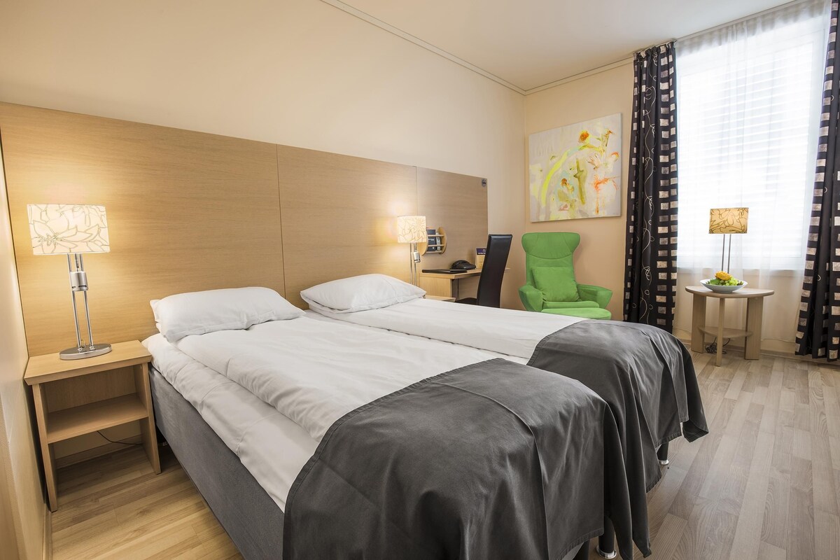 Habitación de hotel con dos camas, un escritorio, una silla, una mesita con un frutero y obras de arte en la pared.