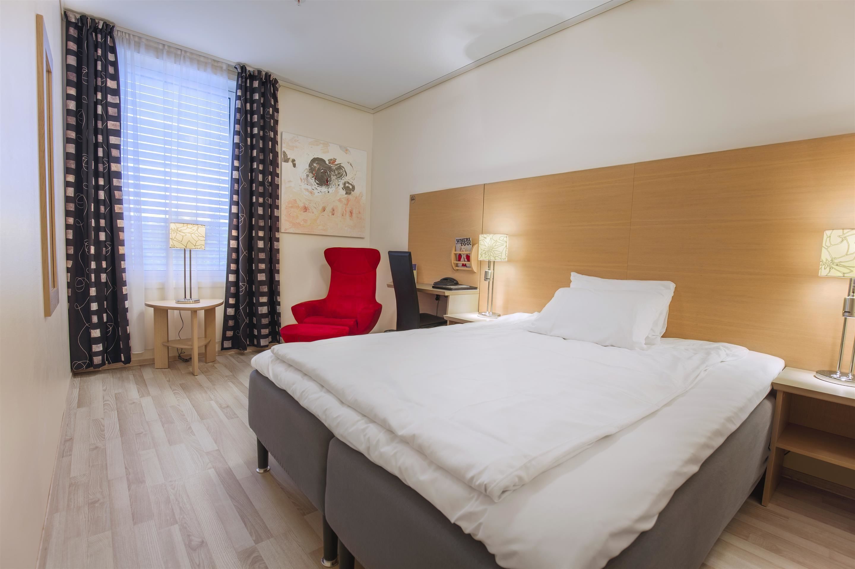 Ett hotellrum med en stor säng, en röd stol, ett litet bord och en vägglampa.