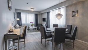 Un área de comedor moderna con una mesa redonda y seis sillas, una mesita auxiliar con una lámpara, y un televisor en la pared.