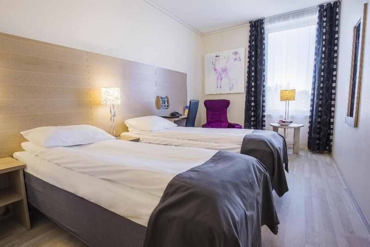 Habitación de hotel con dos camas, una mesita de noche, una silla, una pequeña mesa y una ventana con cortinas.