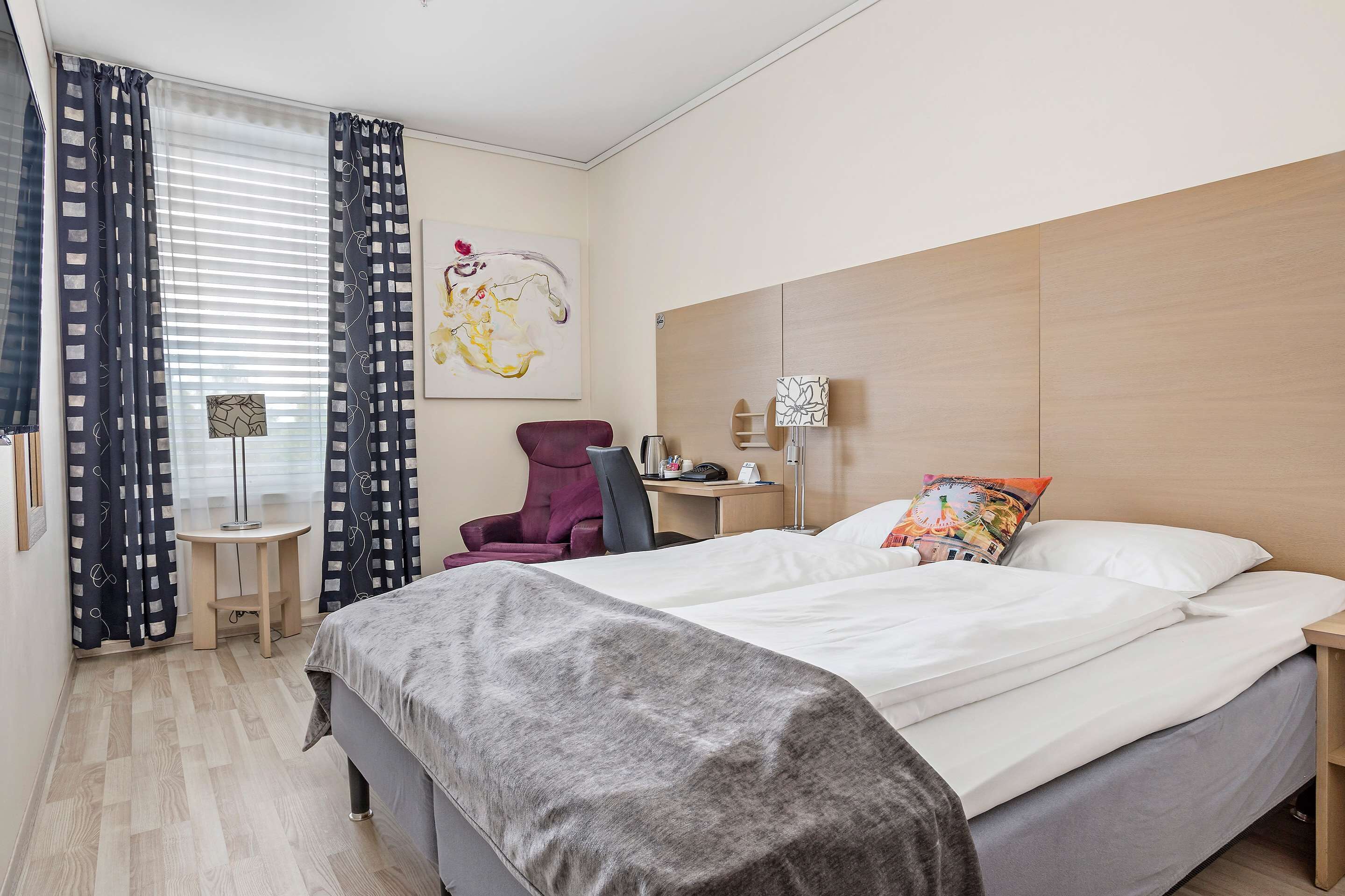 A hotel room with a bed, a desk, a chair, a lamp, and a painting on the wall.