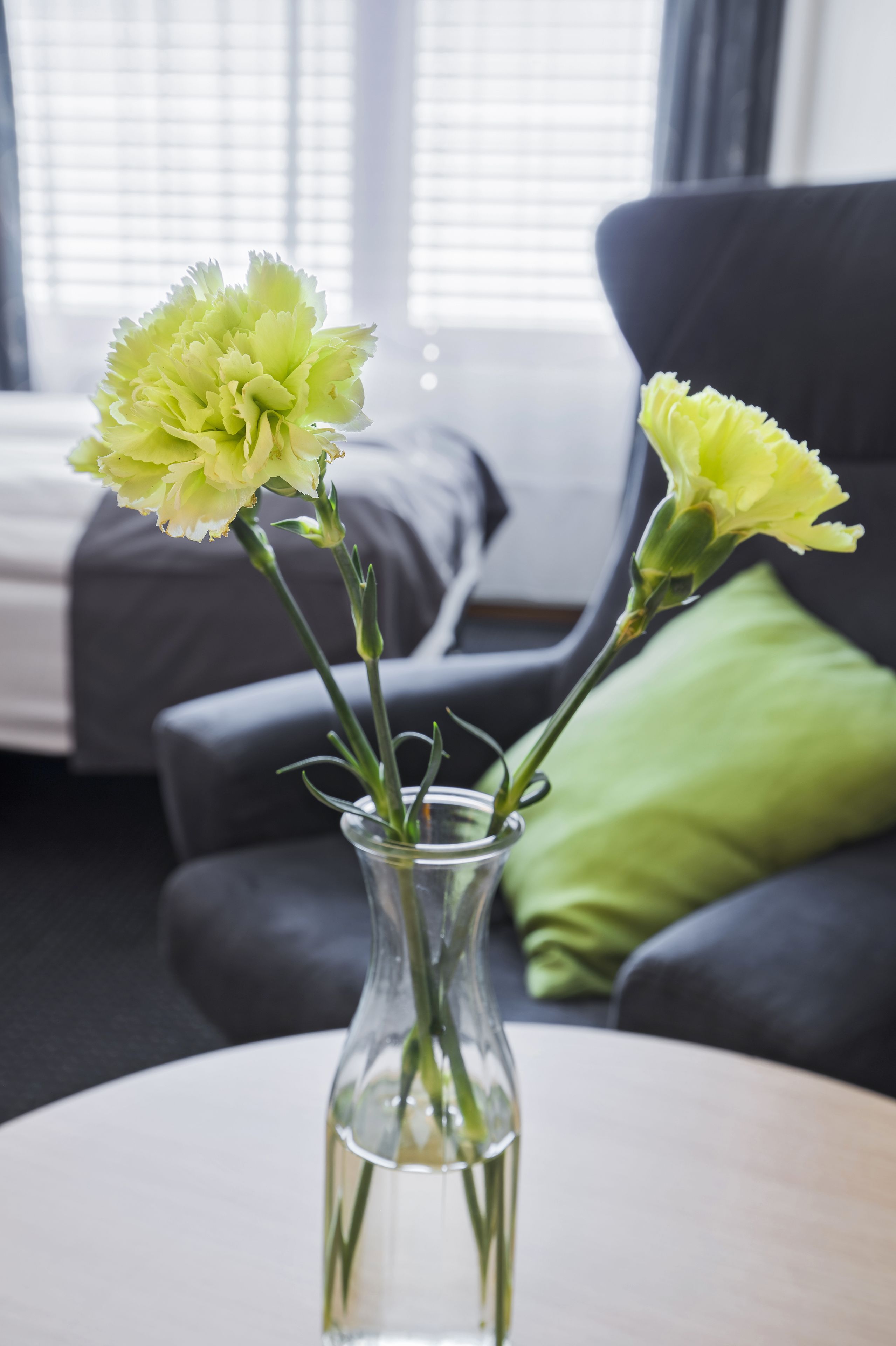 Un vase en verre transparent, orné de fleurs, est posé sur une table ronde dans une pièce où se trouve un lit et, à l’arrière-plan, un coussin vert.