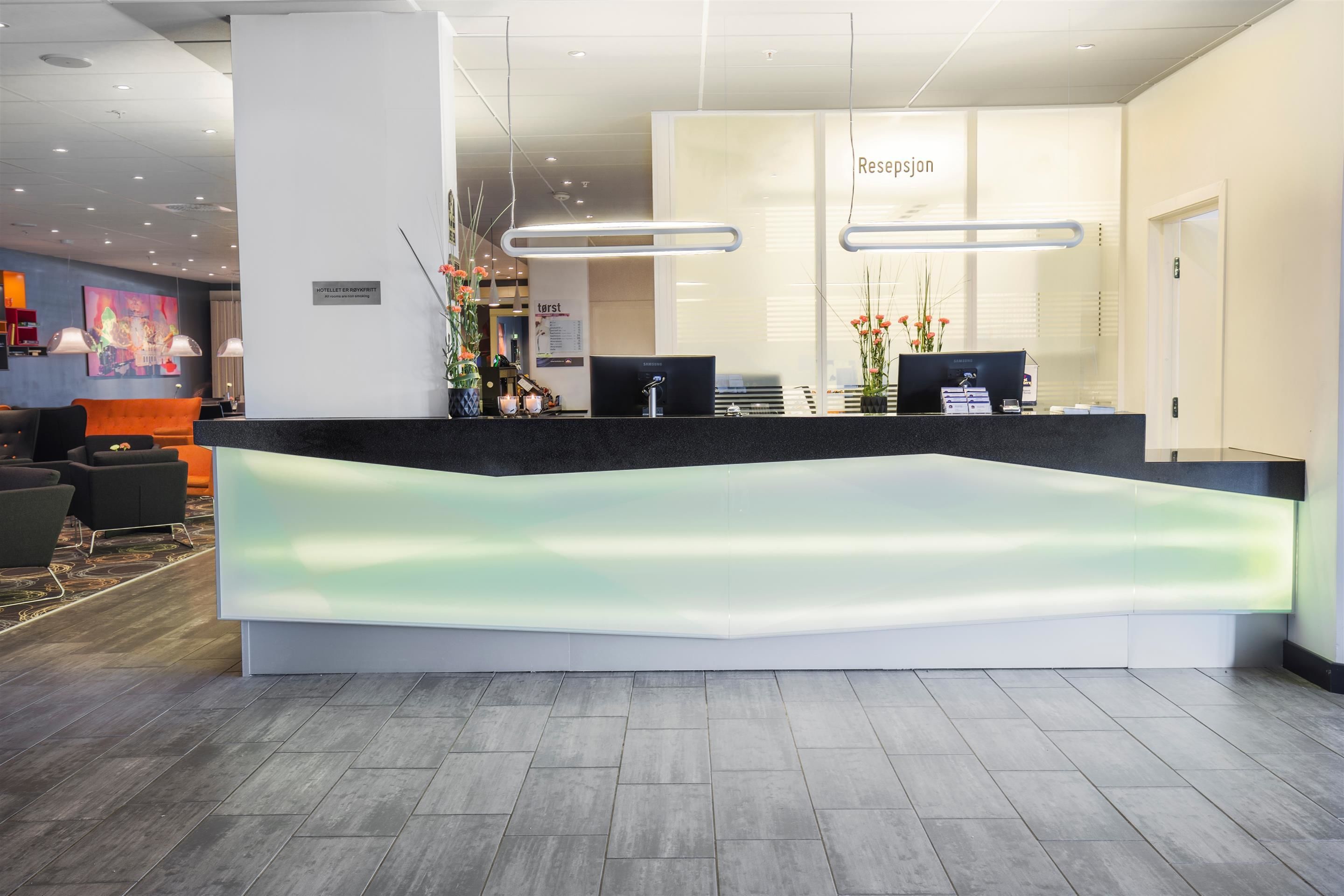 Zona de recepción de hotel con un diseño moderno, que incluye un mostrador de recepción, dos monitores de computadora y un letrero que dice 'Resepsjon'.