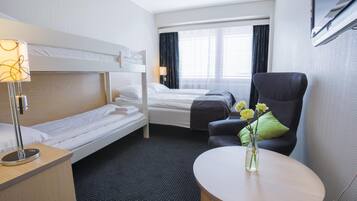 A hotel room with a bunk bed, a single bed, a desk with a lamp, a chair, a small table, and a vase with flowers.