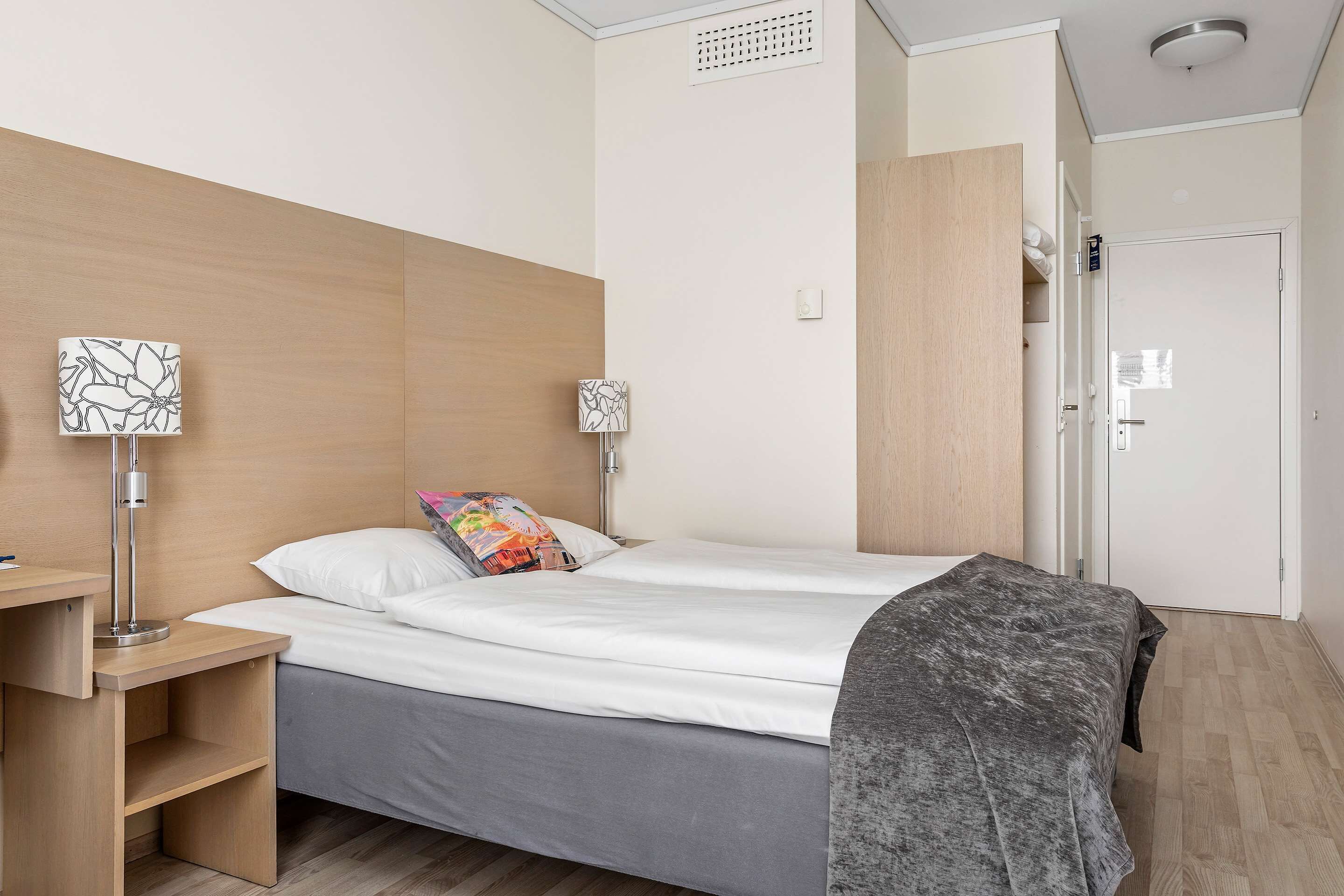 A modern hotel room with a wooden headboard, a bed with white linens, a grey blanket, a bedside table with a lamp, and a wardrobe.
