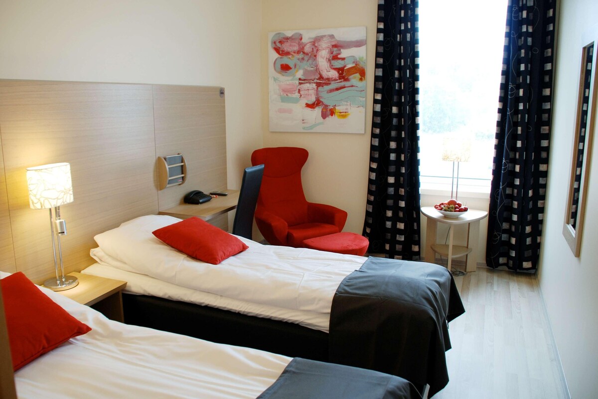 Habitación de hotel con dos camas, un escritorio, una silla, una lámpara, un cuadro y una ventana con cortinas.
