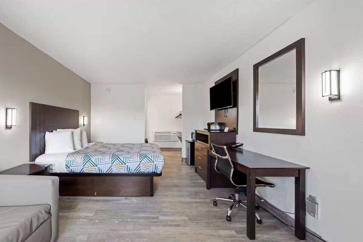 Quarto de hotel com cama, escrivaninha, cadeira, televisão e espelho.