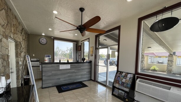 A reception area with a stone wall, a wooden reception desk, a ceiling fan, and a view of the parking lot through glass doors. - Motel 6 Hermison, OR - Umatila (Hermiston)