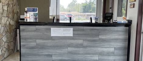 Uma área de recepção com parede de pedra, um relógio, um balcão de atendimento e uma janela com vista para carros estacionados, além de uma placa com medidas de segurança para COVID-19.