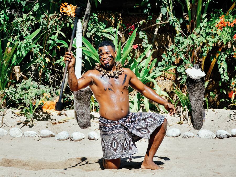 Una persona vestida con traje tradicional sostiene un báculo, de pie sobre arena con plantas y rocas al fondo.
