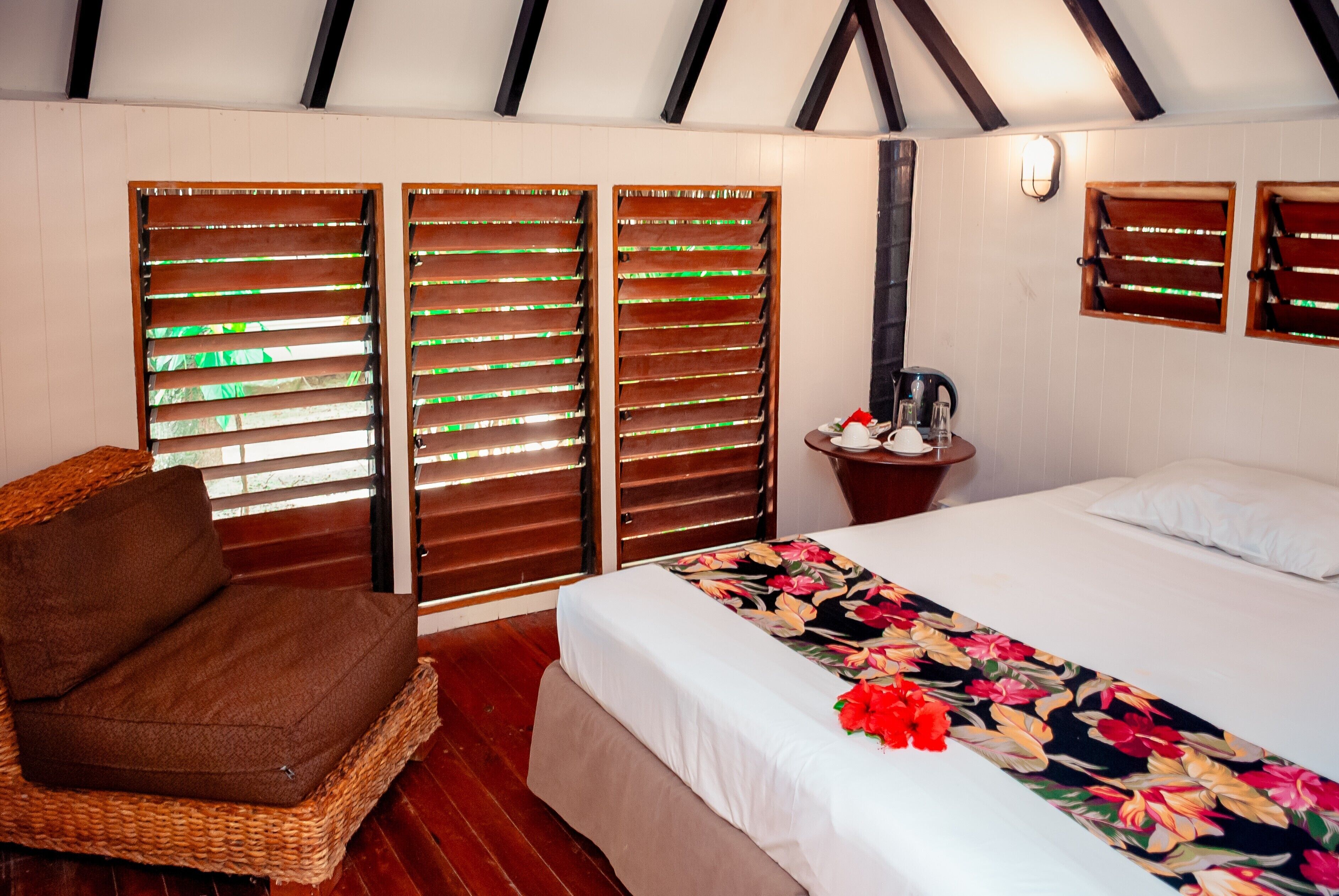 Una habitación con una cama, una silla de mimbre, una mesita de noche con hervidor y contraventanas de madera.