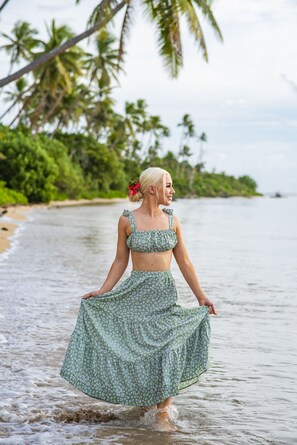 Une femme vêtue d’une robe verte à motifs floraux se promène sur une plage, avec des palmiers en arrière-plan.