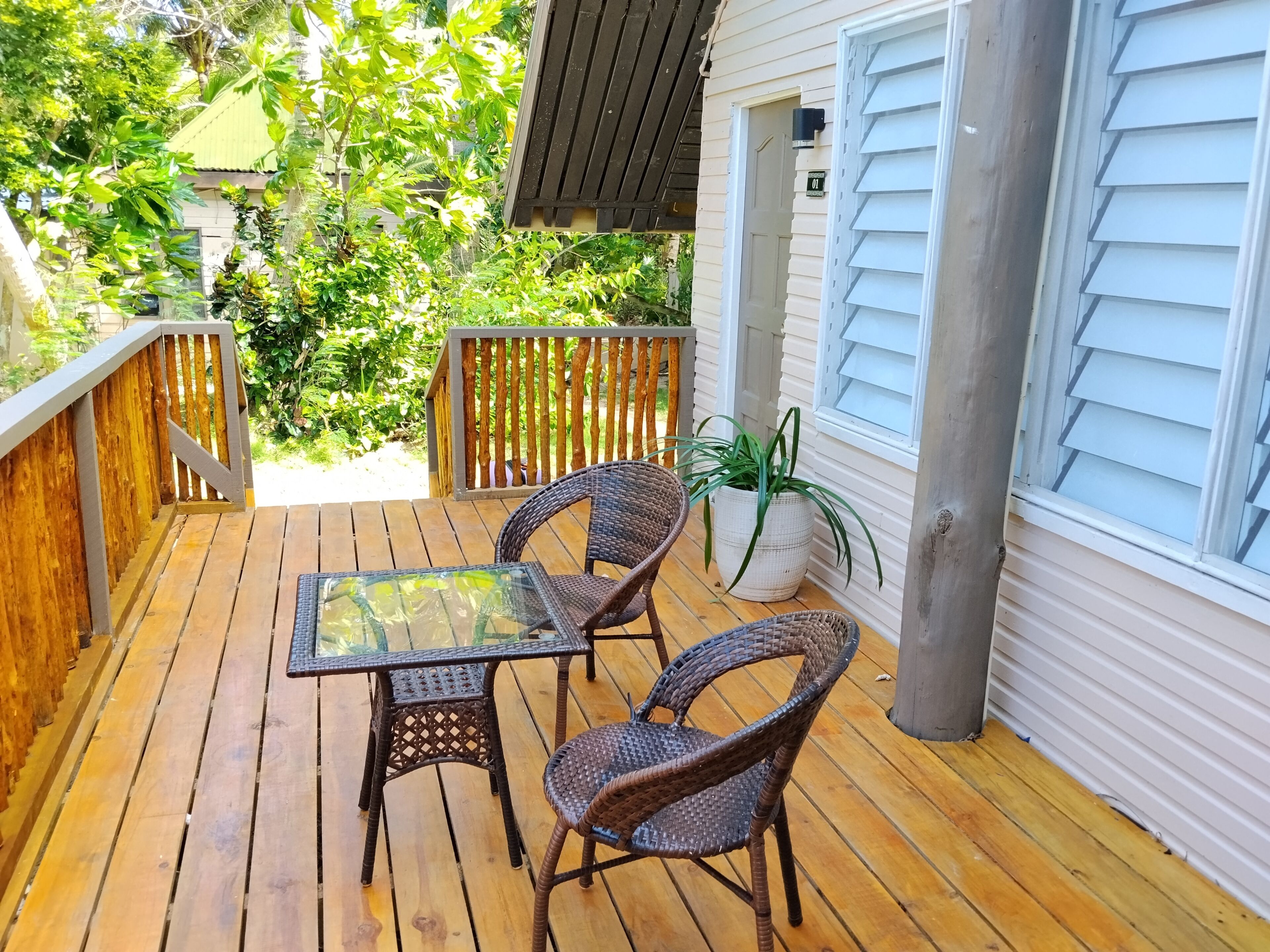 Un patio al aire libre con una mesa de vidrio y sillas de mimbre, rodeado por una terraza de madera y una casa blanca con contraventanas.