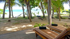 Eine Holzterrasse mit einer gestreiften Liege und einer Hängematte, mit Blick auf einen Strand mit Palmen und dem Meer.