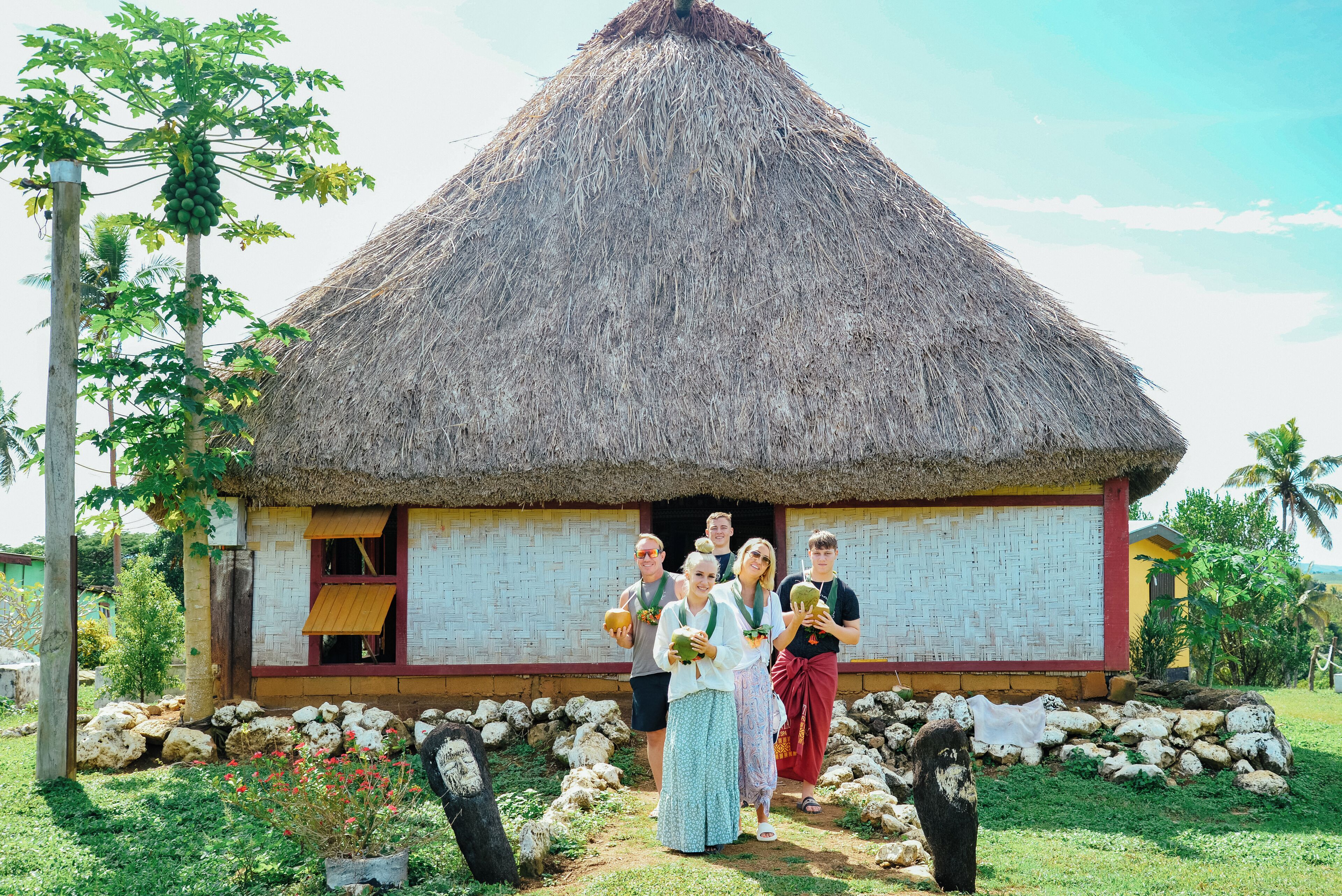 A thatched-roof house with a white exterior and a red base, surrounded by a garden with rocks and a tree.