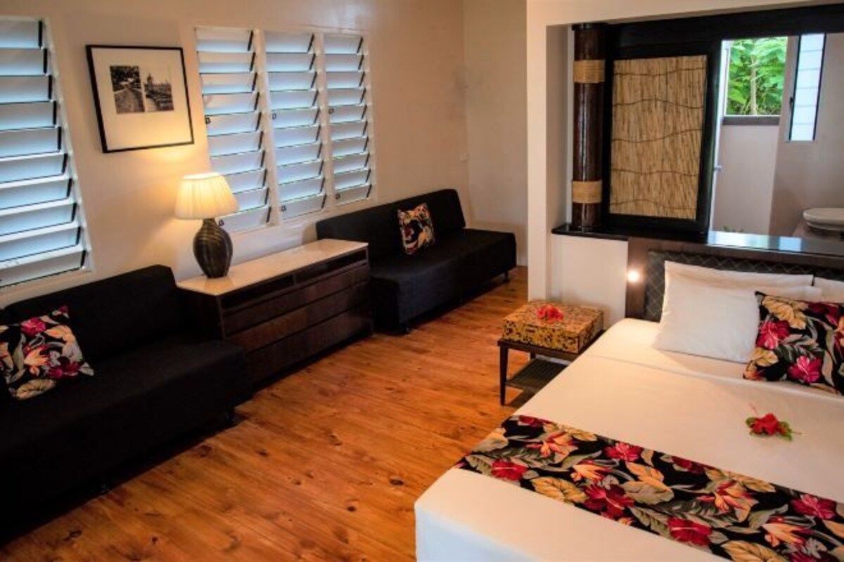 Une chambre d’hôtel avec un lit, un canapé, une petite table et une table de chevet. Il y a une fenêtre avec des stores, un tableau au mur et un miroir au-dessus du lit.