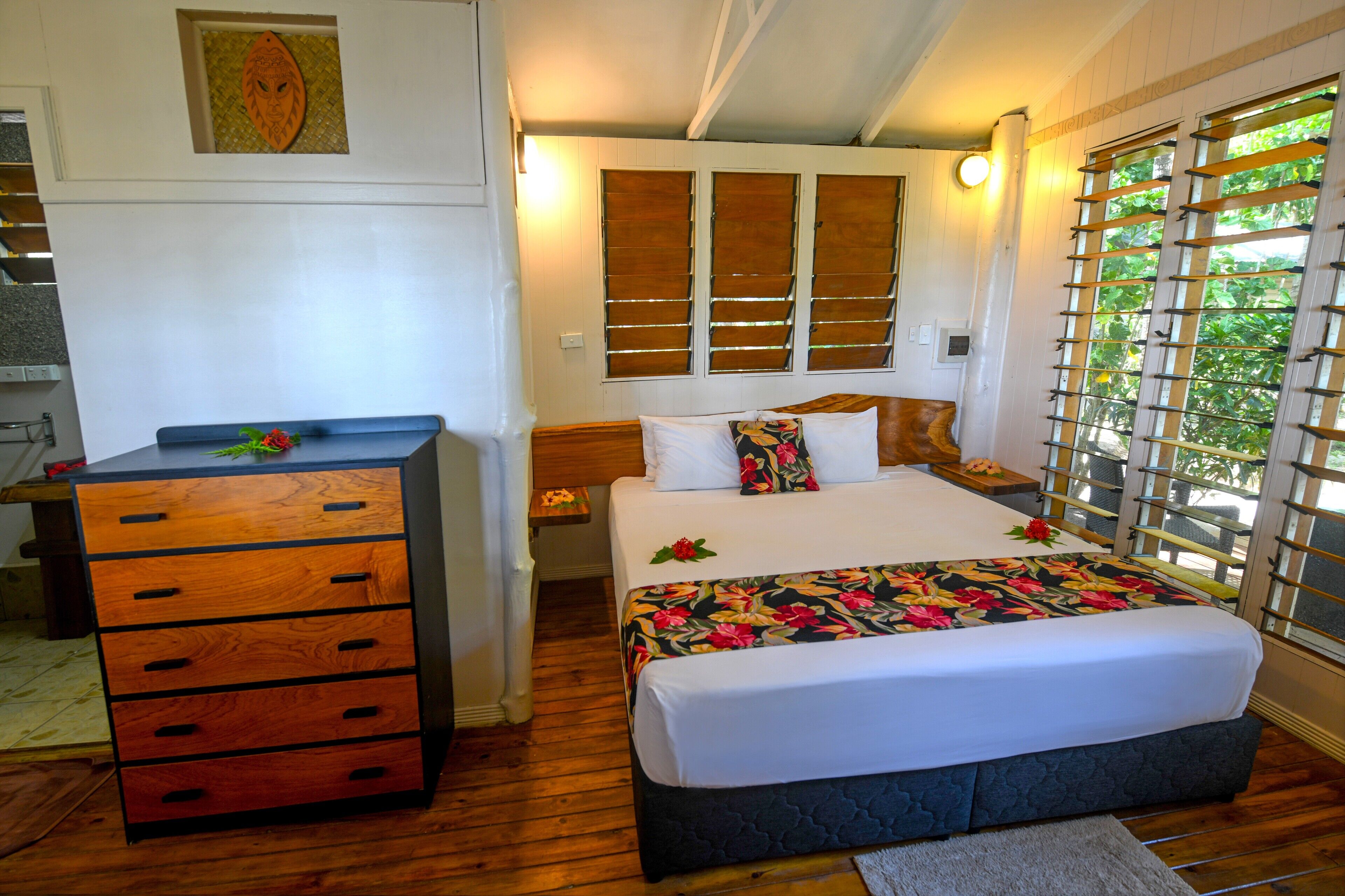 A bedroom with a wooden dresser, a bed with a floral bedspread, and a window with blinds.