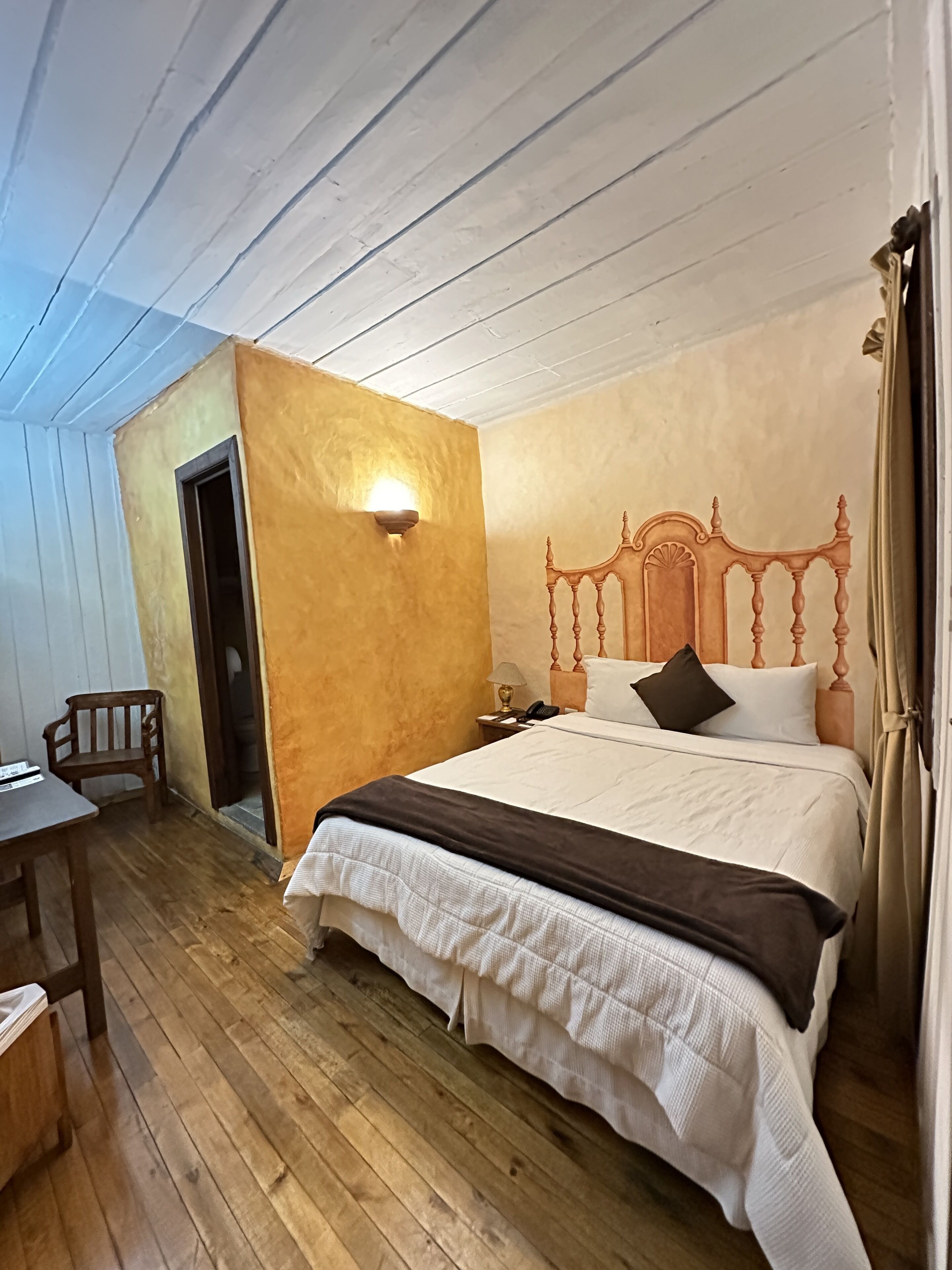 A bedroom with a wooden floor, a bed with a wooden headboard, a nightstand, a chair, and a wall-mounted light fixture.