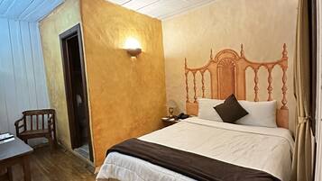A bedroom with a wooden floor, a bed with a wooden headboard, a nightstand, a chair, and a wall-mounted light fixture.