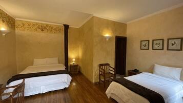 Quarto de hotel com duas camas, piso de madeira, uma pequena cadeira de madeira, mesa de cabeceira com abajur e arte nas paredes.