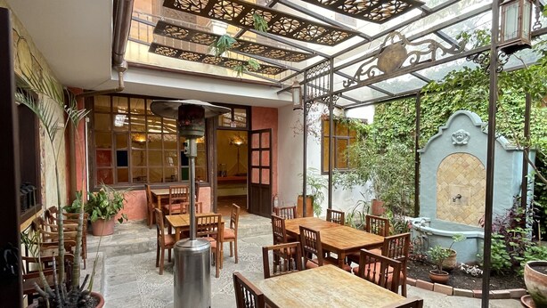 An outdoor patio with wooden tables and chairs, potted plants, and a decorative metal ceiling. - Hotel NASS Casa del Aguila (Cuenca)