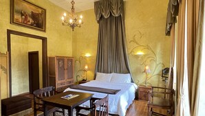 A hotel room with a wooden floor, a bed with white linens, a wooden table with chairs, a chandelier, and a painting on the wall. - Hotel NASS Casa del Aguila (Cuenca)