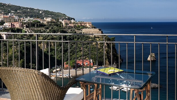 A balcony with wicker furniture, a glass table, and a view of the sea and a town in the background. - Villa Ketty (Vico Equense)