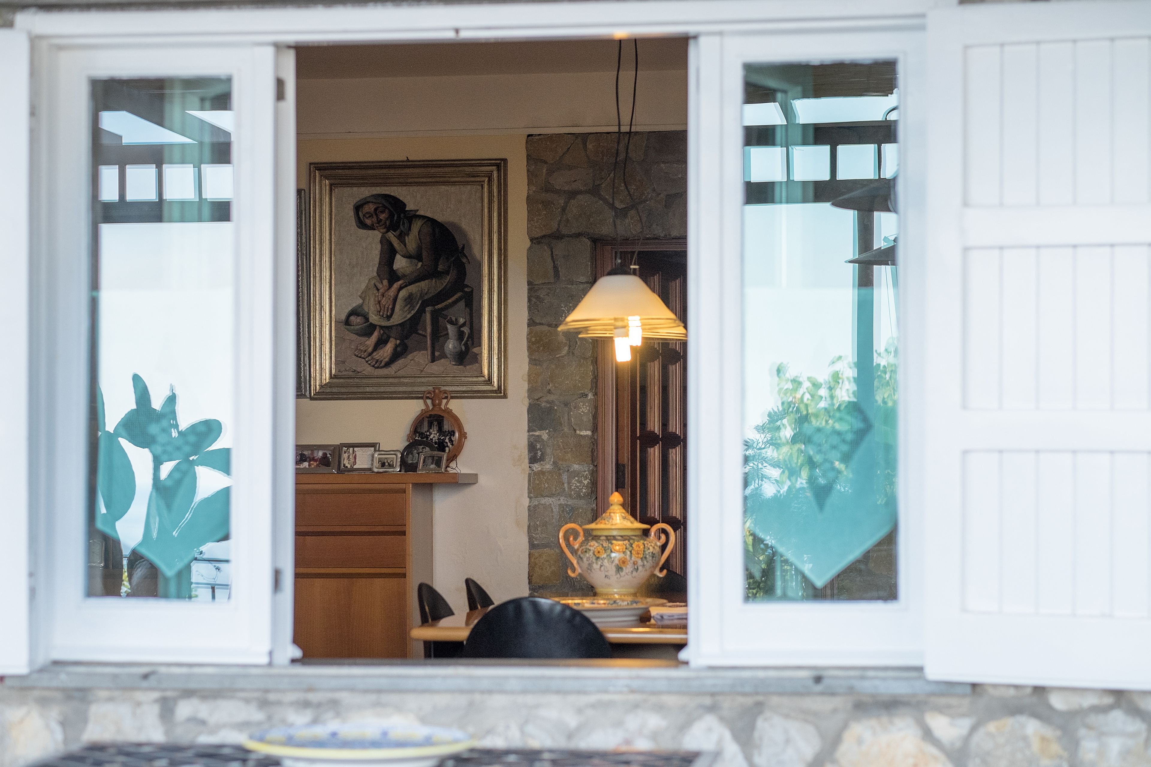 A room with a stone wall, a wooden cabinet, a table with chairs, a lamp, and a framed picture.