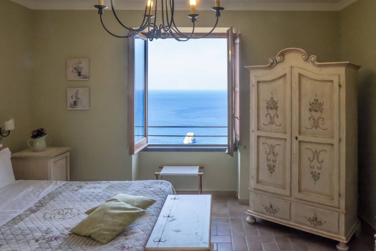 Ein Schlafzimmer mit Bett, Nachttisch, Kommode, einem Fenster mit Meerblick und einer Chandelier.