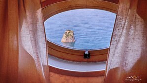 A view from inside a room with a wooden window frame and curtains, looking out at a calm body of water with a small rock formation. - Villa Ketty (Vico Equense)