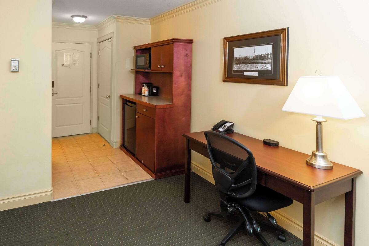 A hotel room with a wooden desk, a chair, a telephone, a lamp, a microwave, and a framed picture on the wall.