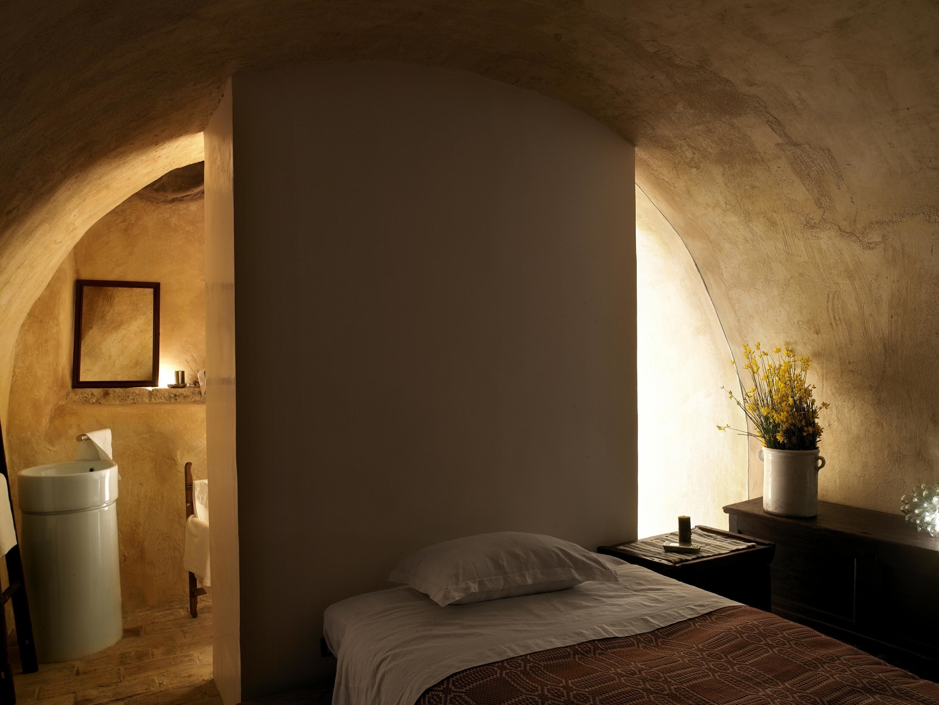 A bedroom with a bed, a nightstand, a potted plant, a mirror, and a sink in a stone-walled alcove.