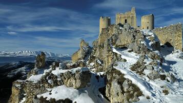 Un castillo de piedra histórico con múltiples torres, rodeado de terreno rocoso cubierto de nieve y montañas al fondo.