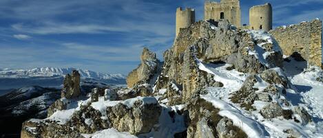 Un castillo de piedra histórico con múltiples torres, rodeado de terreno rocoso cubierto de nieve y montañas al fondo.