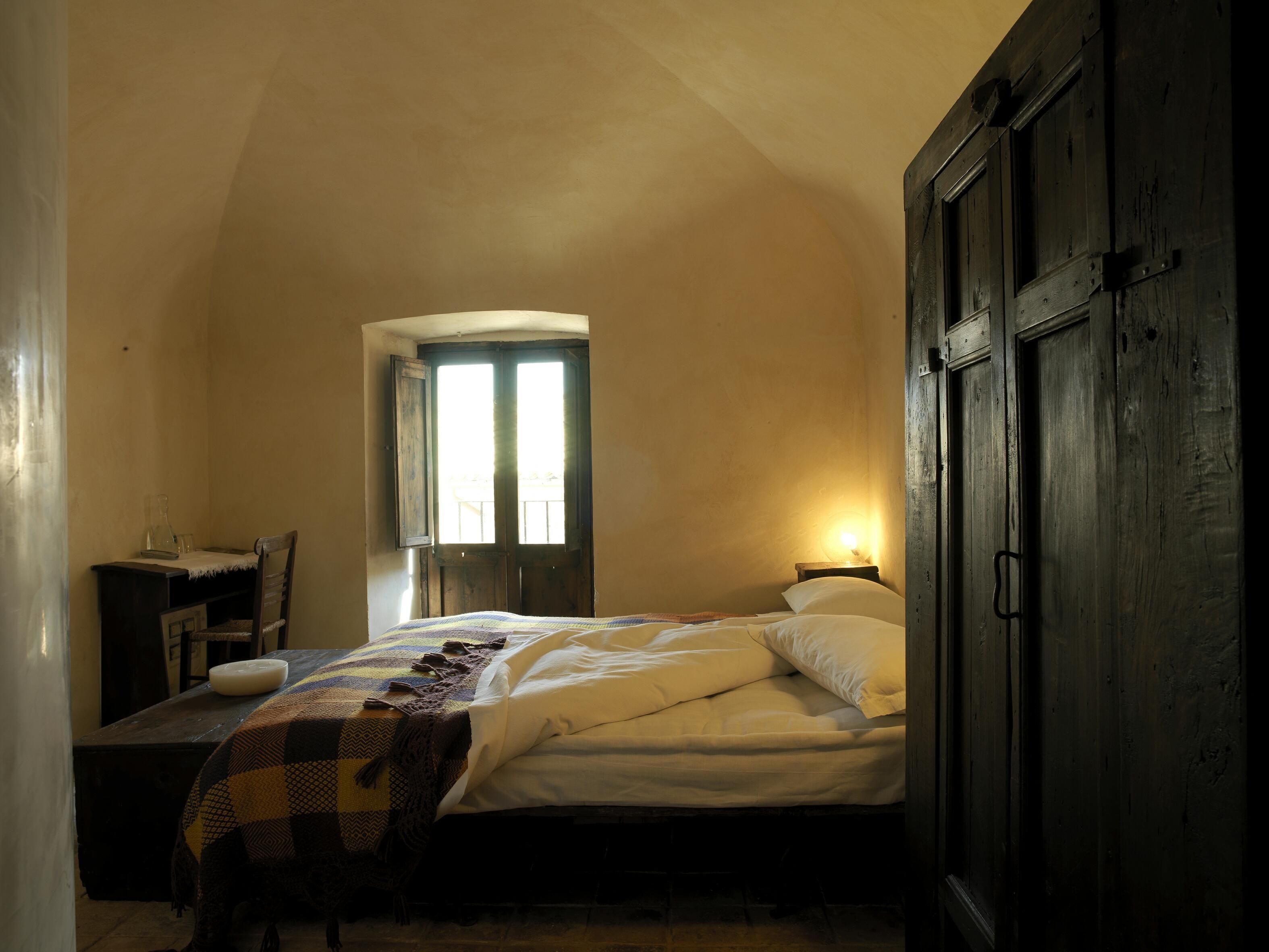 A bedroom with a bed, a desk, and a chair. There is a window with a view of the outside.