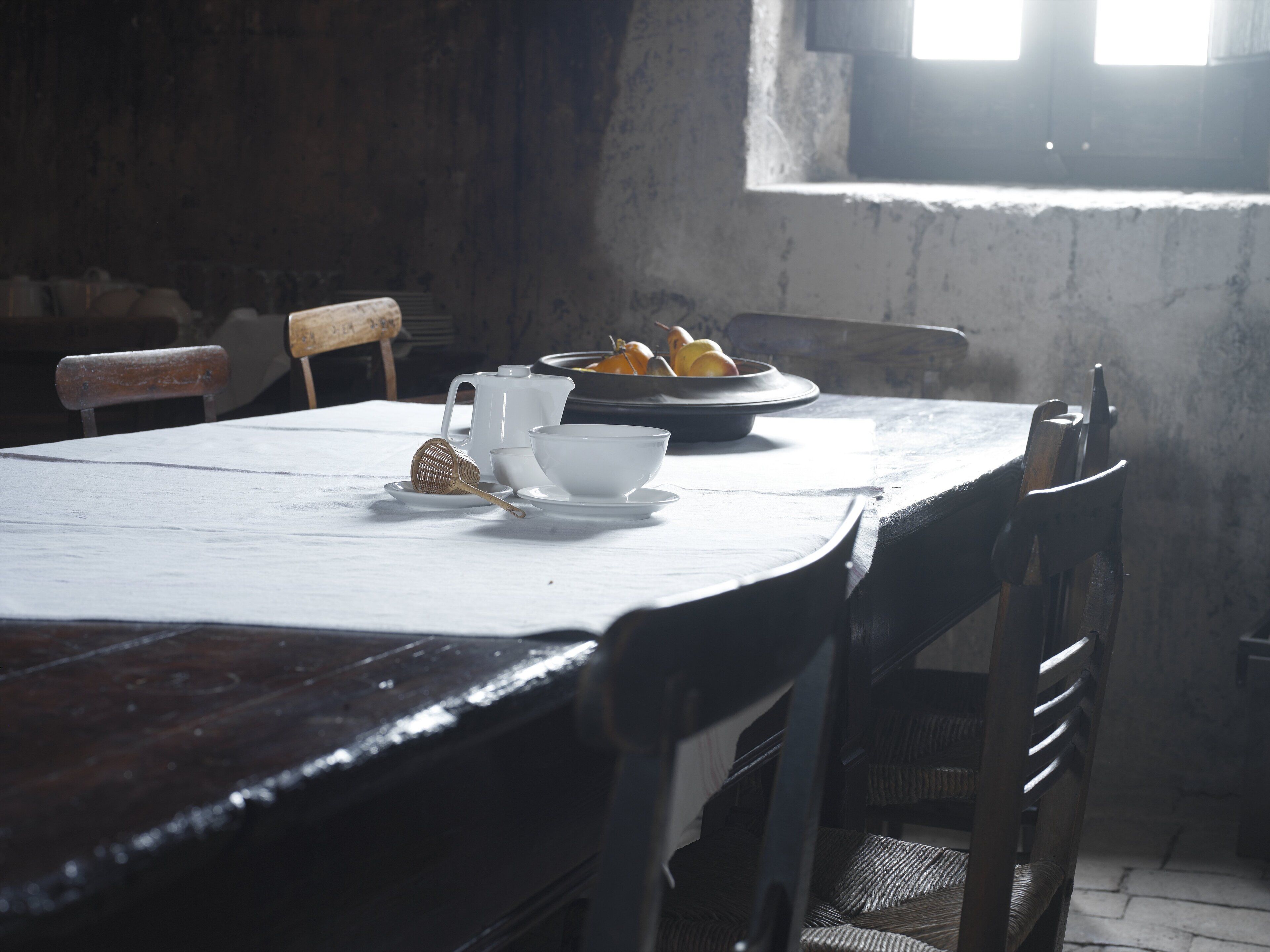 A dining table with a white tablecloth, a bowl of fruit, and a teapot.