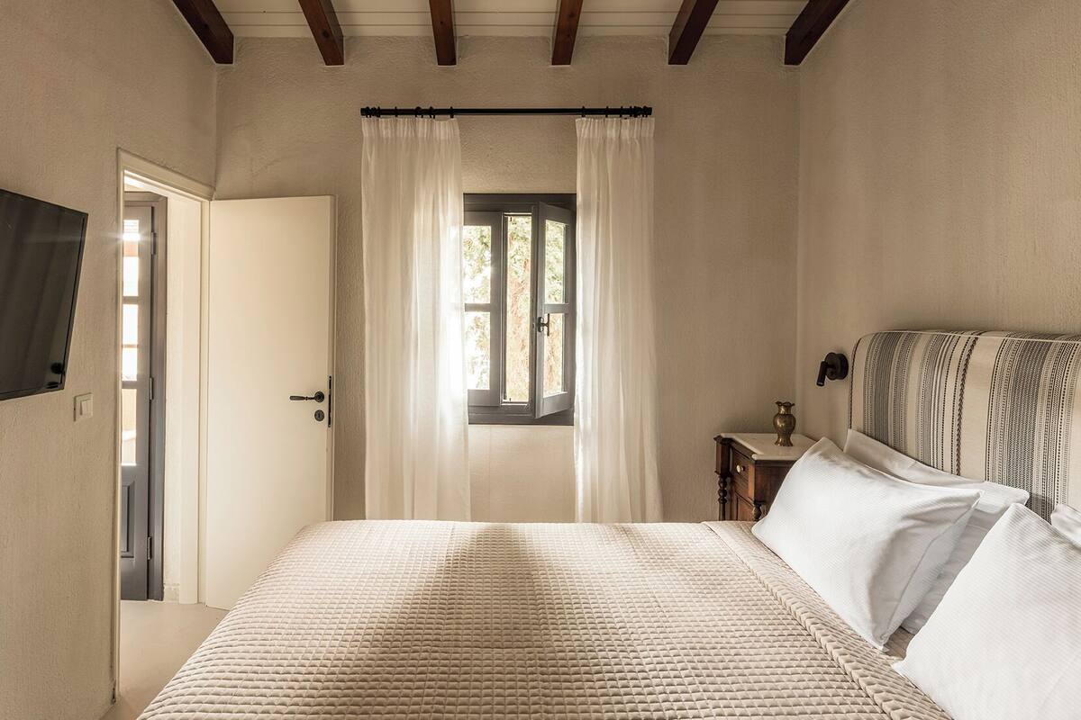 Une chambre à coucher avec un lit, une fenêtre avec des rideaux, une table de chevet avec une lampe et un téléviseur fixé au mur.