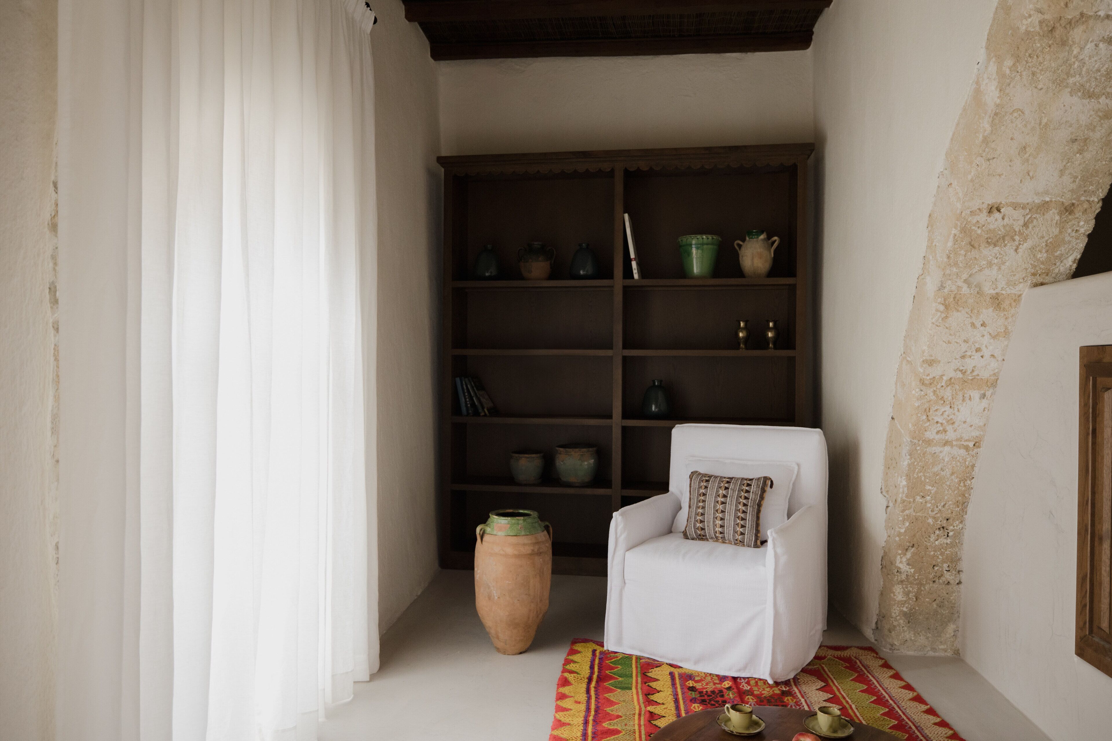 Una habitación con una silla blanca, una estantería con cerámica y un jarrón grande.