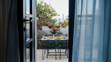 An outdoor dining area with a table set for two, surrounded by greenery and a white wall.