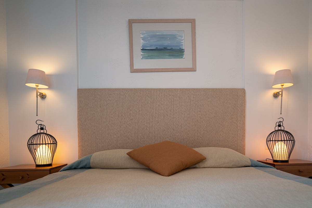 Un lit bien fait, avec un seul oreiller marron, deux lampes fixées au mur et un tableau encadré au-dessus de la tête de lit.