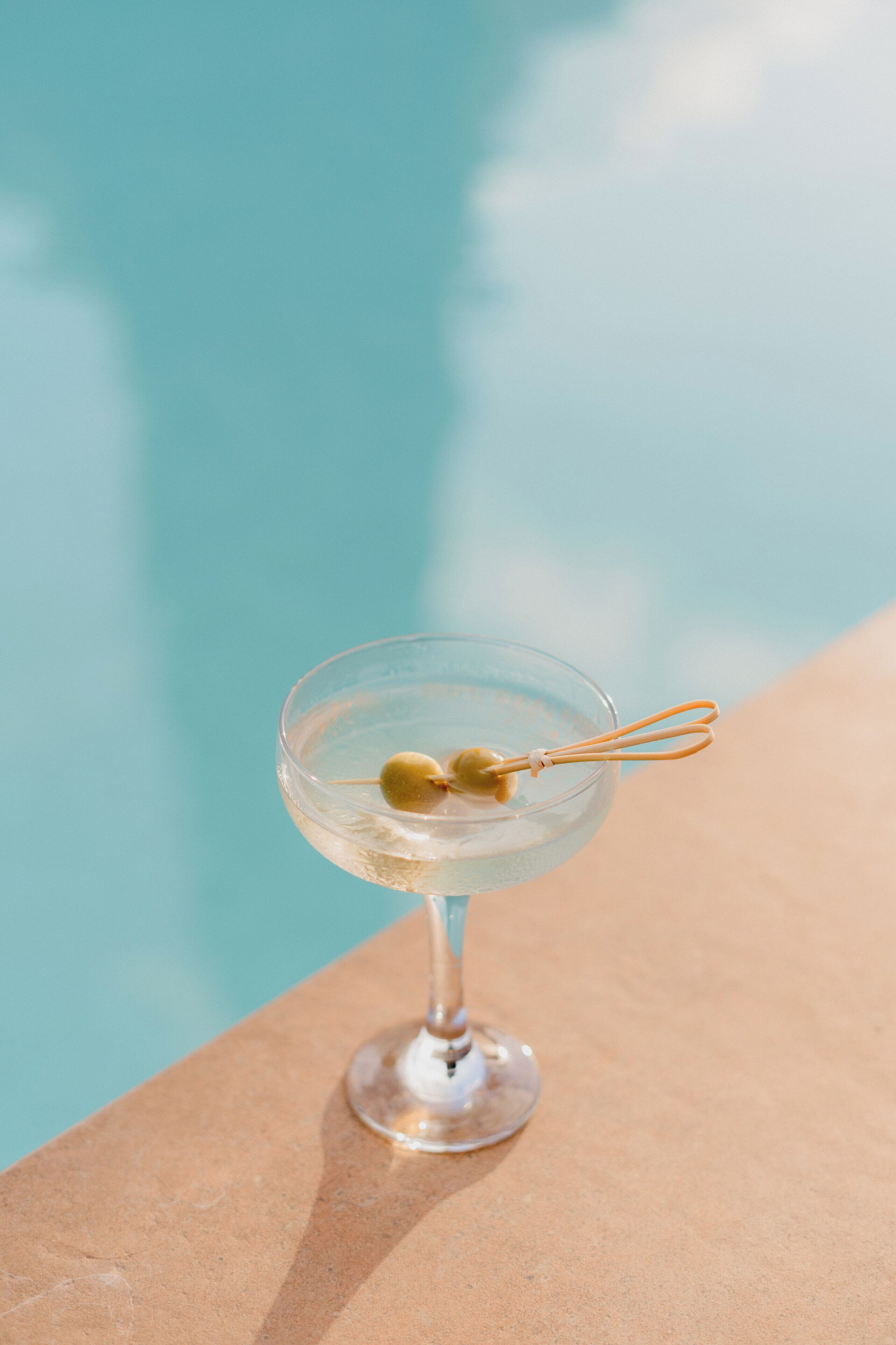 A clear glass cocktail with an olive garnish, placed on a poolside ledge.