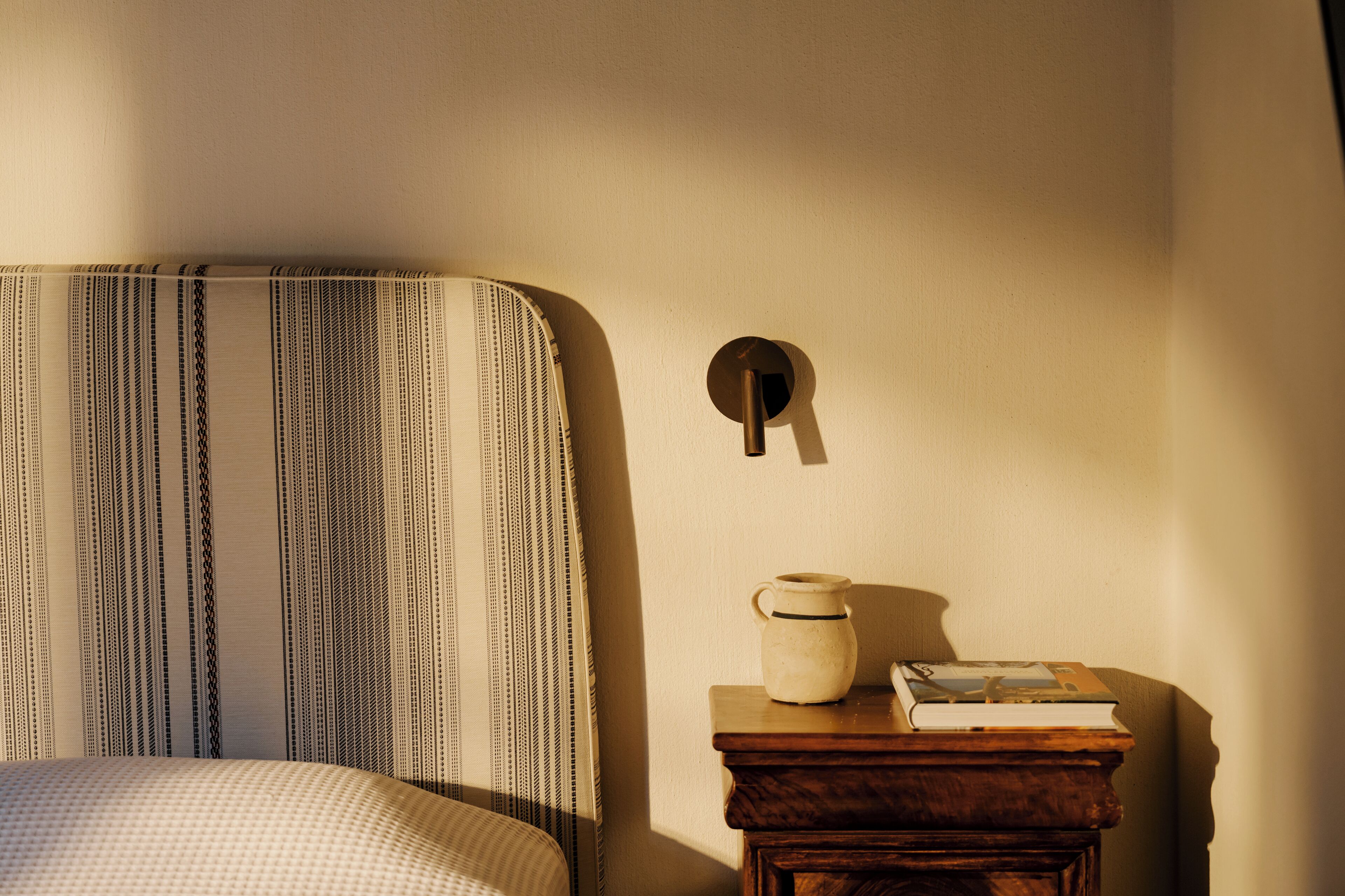 Una cama con ropa de cama a rayas, una mesita de noche con un libro y un florero, y una lámpara de pared.