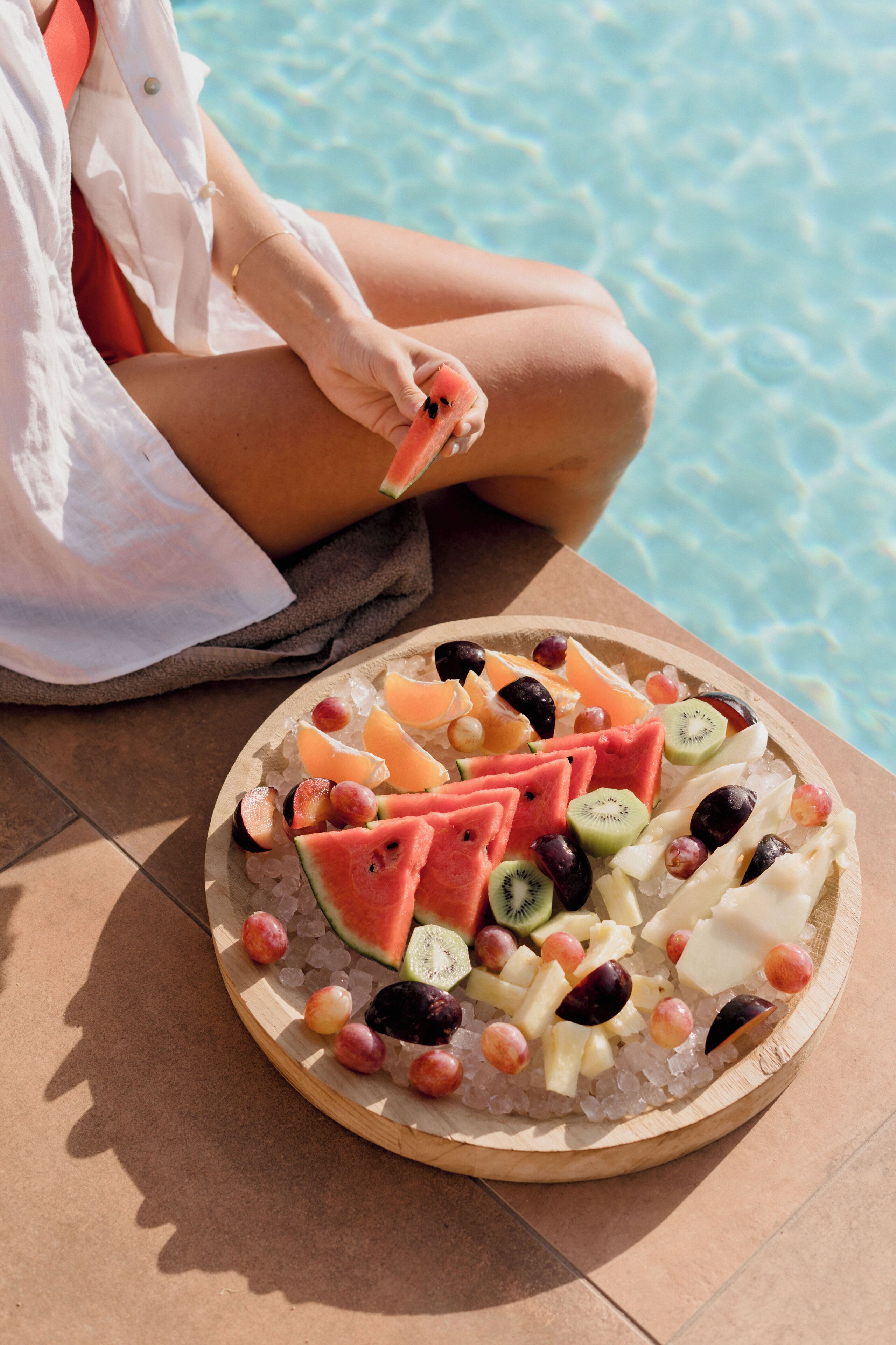 Una bandeja redonda con fruta, sandía, kiwi, uvas y queso sobre una superficie de baldosas cerca de una piscina.
