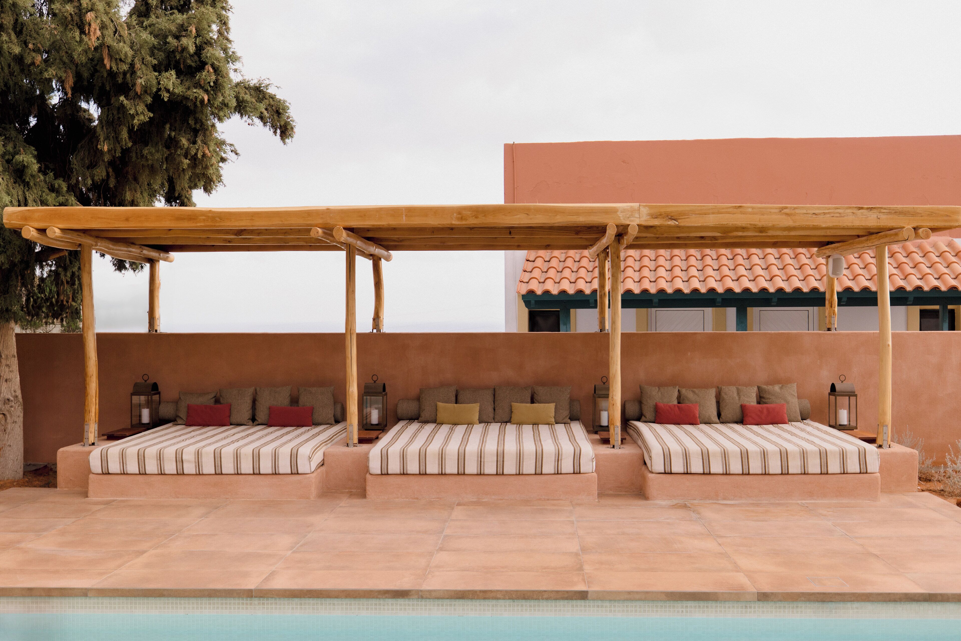 A poolside area with a covered seating area, a bed with striped bedding, and a view of a building with a tiled roof.