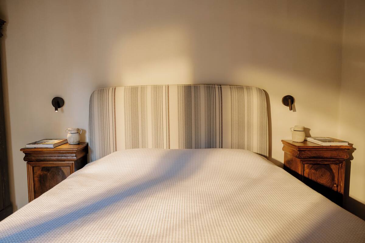 Un lit bien fait, avec une tête de lit à rayures, deux tables de chevet dotées de lampes et un petit objet décoratif sur chacune.