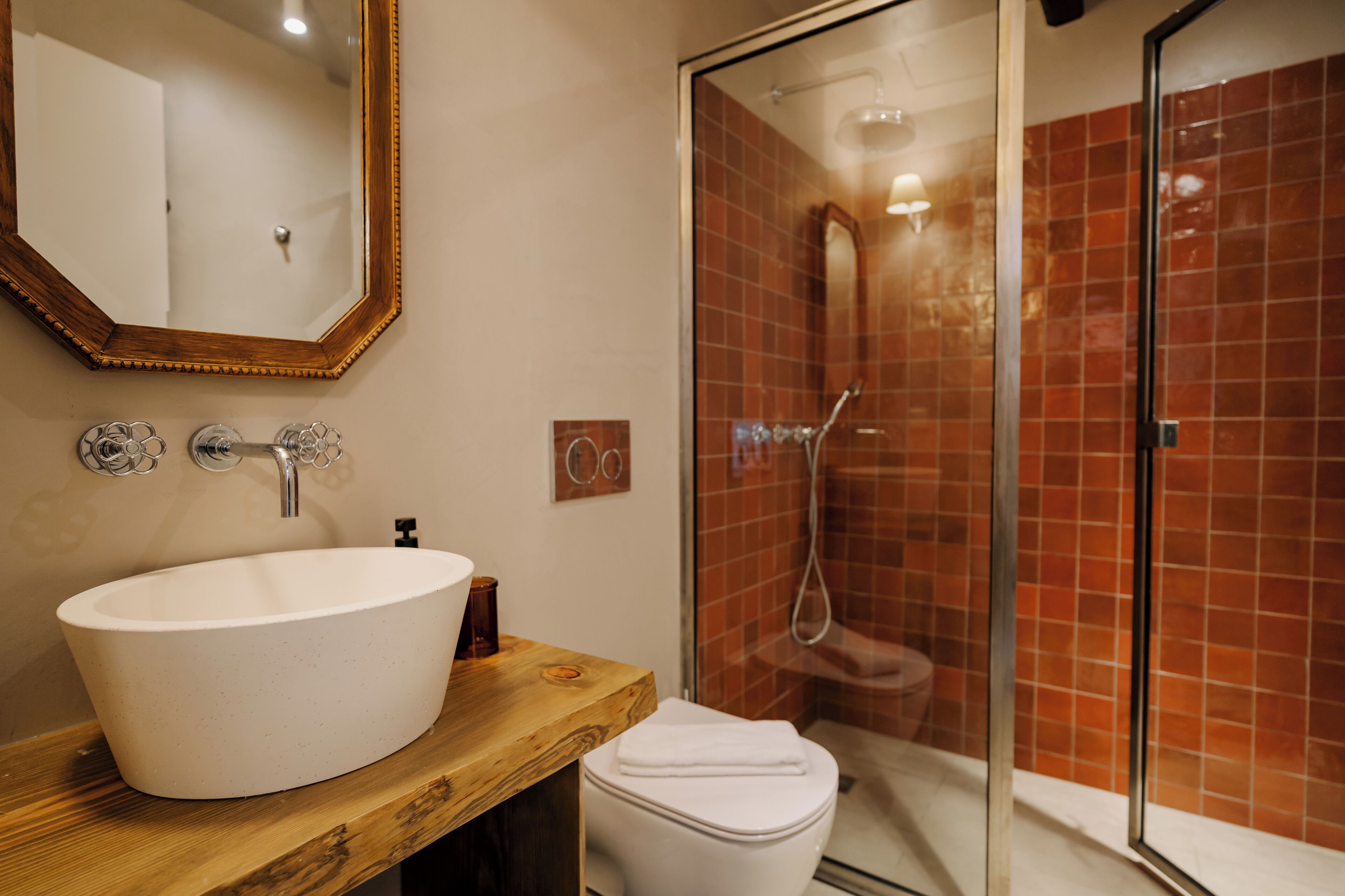 Une salle de bain avec un meuble-lavabo en bois, un lavabo blanc, un miroir et une douche en verre avec des carreaux rouges.
