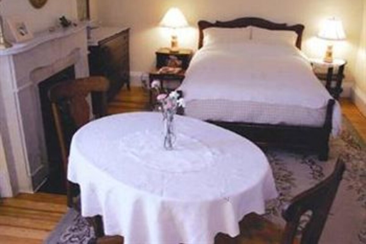 A bedroom with a bed, bedside lamps, a round table with a vase, and a fireplace.
