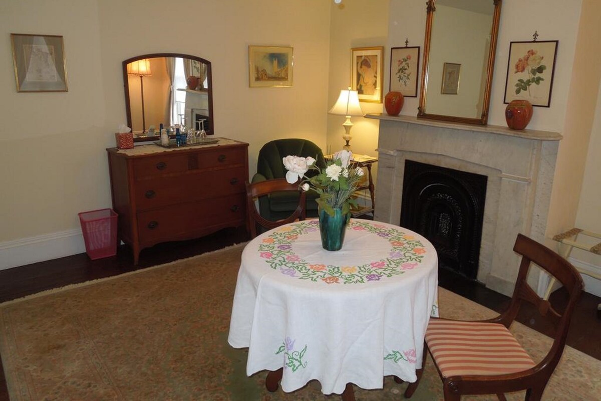 A cozy room with a fireplace, a round table with a floral tablecloth, a wooden dresser, and a mirror on the wall.